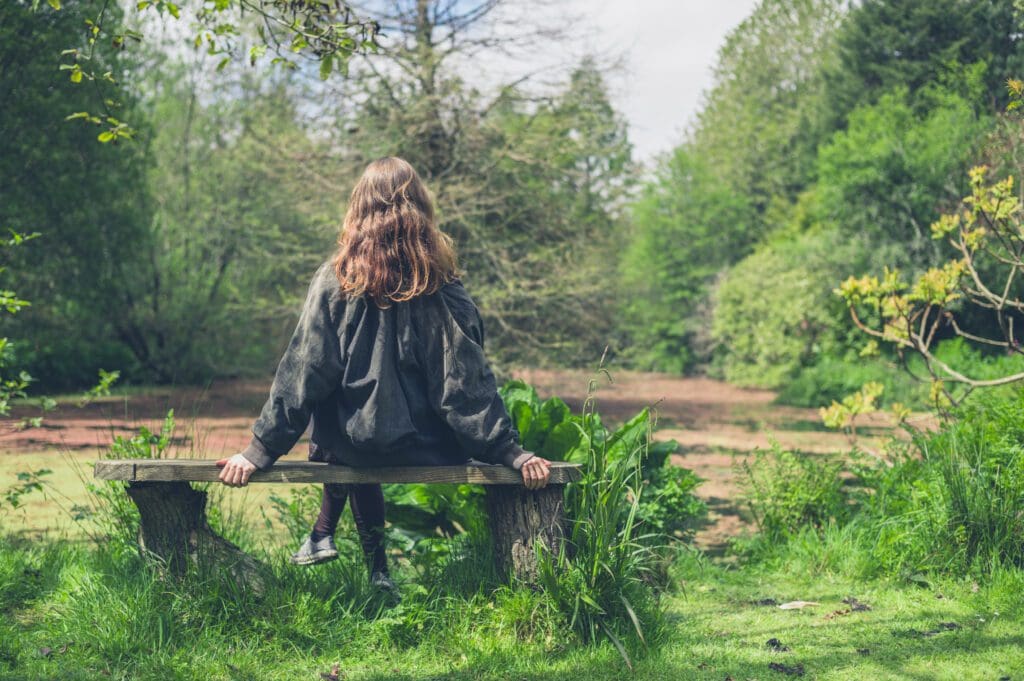 A person sits on a bench outside in the sun, surrounded by nature and greenery.